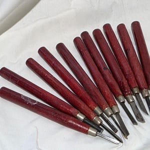 May include: A set of ten carving tools with cylindrical dark red wooden handles and silver-toned metal ferrules. Each tool features a distinct metal blade tip, including various gouge and chisel shapes, arranged in a diagonal fan formation.