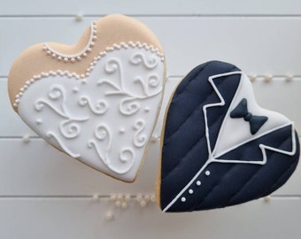 May include: Two heart-shaped cookies decorated with icing to resemble a bride and groom. The bride cookie is light brown with white icing and the groom cookie is dark blue with white icing.