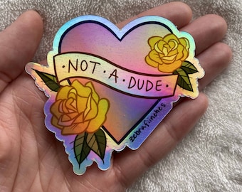 May include: A holographic sticker with a heart shape and two yellow roses. The heart has a banner that reads "NOT. A. DUDE." The sticker is made by Zebrafinches.