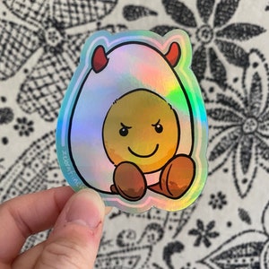 Op de afbeelding: Een holografische sticker met een cartoon-ei met duivelshoorns en een ondeugende glimlach. Het ei draagt ​​een witte schaal met een regenboogkleurige, iriserende glans.