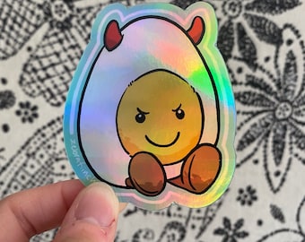 Pode incluir: Um adesivo holográfico de um ovo de desenho animado com chifres de diabo e um sorriso travesso. O ovo está usando uma casca branca com um brilho iridescente arco-íris.