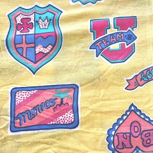 Può includere: Un tessuto giallo con toppe colorate. Le toppe presentano uno scudo con una corona, una lettera U con la parola "TEAM" su uno stendardo, un rettangolo con la parola "Monaco" e una palma, e un cuore con la parola "No. 8" all'interno.