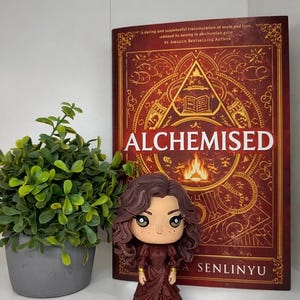 Może przedstawiać: Figurka Funko Pop kobiety z brązowymi włosami i bordową sukienką stoi przed książką zatytułowaną "ALCHEMISED" autorstwa A. Senlinyu. Książka ma czerwoną okładkę ze złotymi akcentami i tekstem. Po lewej stronie znajduje się mała roślina doniczkowa.