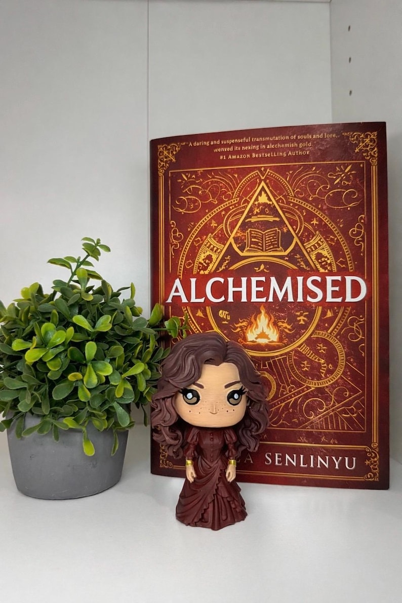Może przedstawiać: Figurka Funko Pop kobiety z brązowymi włosami i bordową sukienką stoi przed książką zatytułowaną "ALCHEMISED" autorstwa A. Senlinyu. Książka ma czerwoną okładkę ze złotymi akcentami i tekstem. Po lewej stronie znajduje się mała roślina doniczkowa.