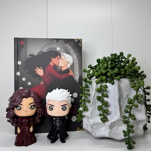 Może przedstawiać: Kompozycja z dwiema figurkami Funko Pop, książką i białym ceramicznym pojemnikiem na rośliny z wiszącą zieloną rośliną. Figurki przedstawiają postacie z książki. Okładka książki przedstawia dwie osoby obejmujące się. Doniczka ma unikalny, teksturowany design.