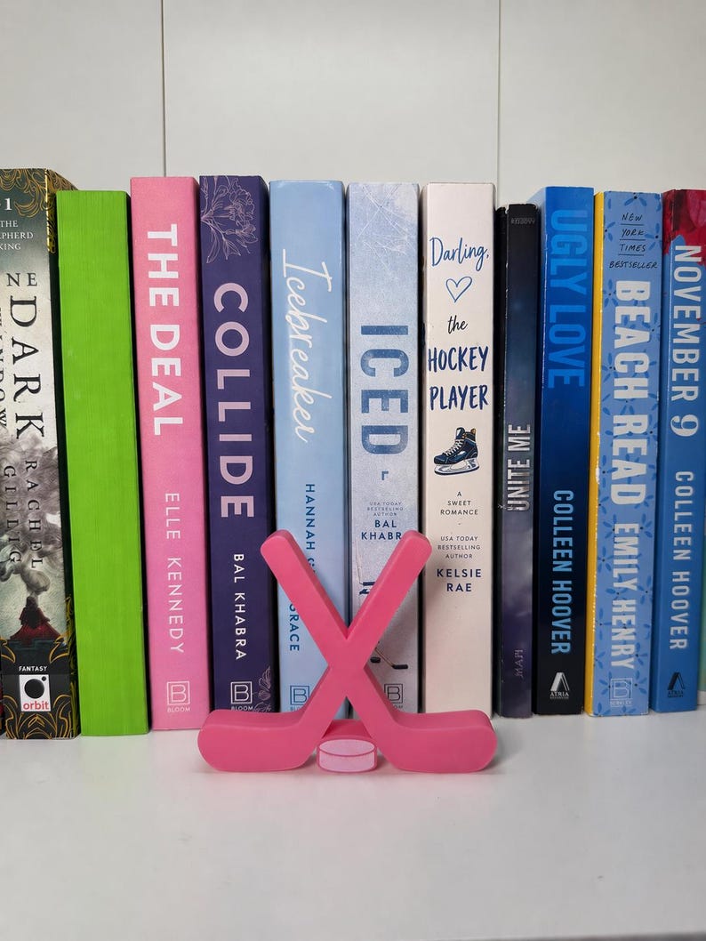 May include: A collection of books with colorful spines, including titles like "The Deal" and "Ugly Love." A pink hockey stick and puck bookend sits in front of the books. The books are arranged on a white shelf.