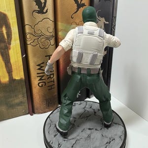 May include: A figurine of a soldier in a green balaclava, green pants, and a white vest. The figure is standing on a gray, textured circular base. The figure is posed with arms outstretched. Books are in the background.