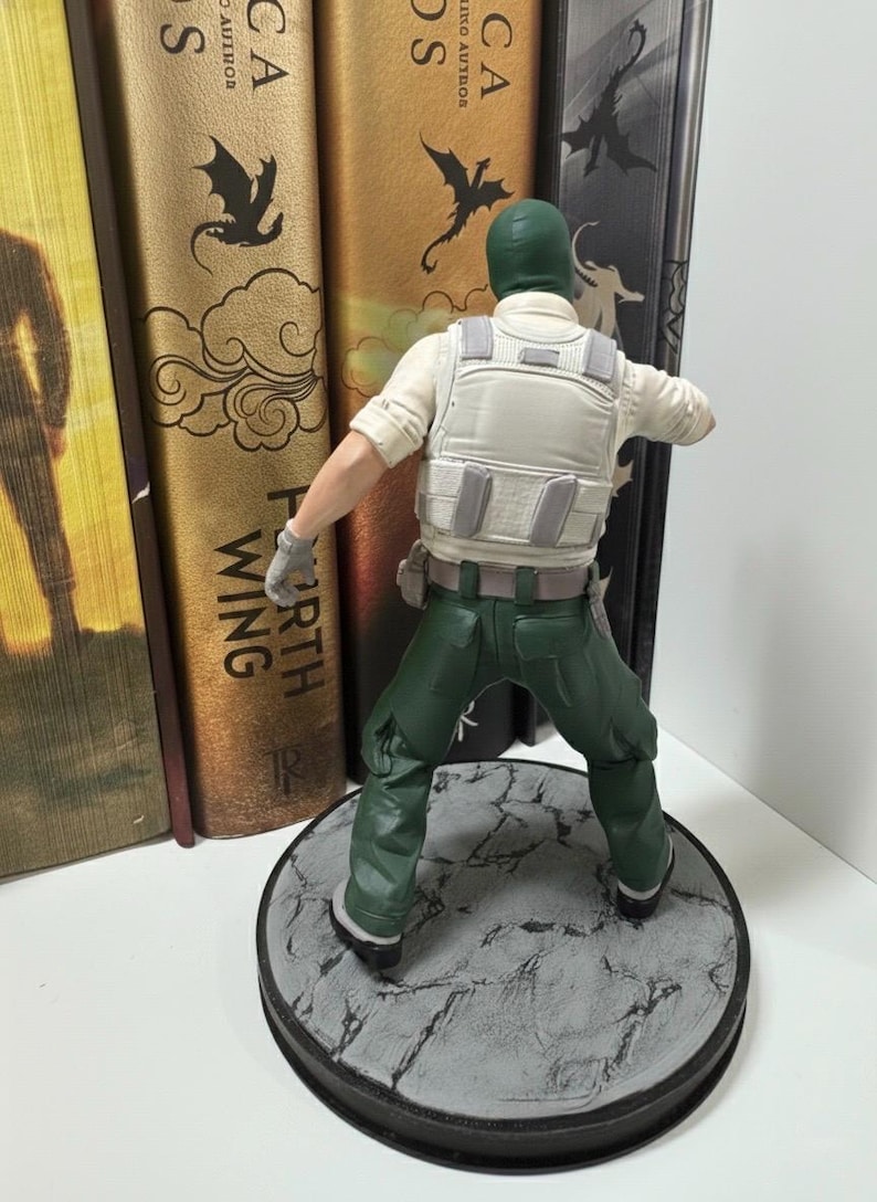 May include: A figurine of a soldier in a green balaclava, green pants, and a white vest. The figure is standing on a gray, textured circular base. The figure is posed with arms outstretched. Books are in the background.