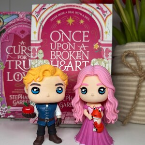 K&ouml;nnte beinhalten: Zwei Funko Pop-Figuren von Charakteren aus einem Fantasy-Roman. Eine Figur hat blondes Haar, eine blaue Weste und h&auml;lt einen roten Apfel. Die andere Figur hat rosa Haare, ein rosa Kleid und h&auml;lt einen kleinen orangefarbenen Fuchs. Das Buchcover zeigt "Once Upon a Broken Heart."