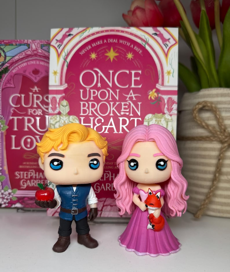 K&ouml;nnte beinhalten: Zwei Funko Pop-Figuren von Charakteren aus einem Fantasy-Roman. Eine Figur hat blondes Haar, eine blaue Weste und h&auml;lt einen roten Apfel. Die andere Figur hat rosa Haare, ein rosa Kleid und h&auml;lt einen kleinen orangefarbenen Fuchs. Das Buchcover zeigt "Once Upon a Broken Heart."