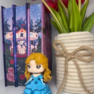 May include: A figurine of a woman with blonde hair and a blue dress stands in front of a row of books. The books have a carousel design. A vase with red tulips and green leaves is to the right.