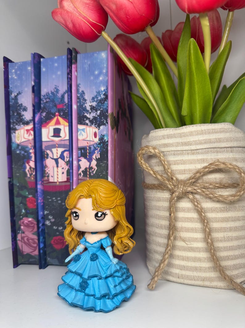 May include: A figurine of a woman with blonde hair and a blue dress stands in front of a row of books. The books have a carousel design. A vase with red tulips and green leaves is to the right.