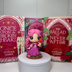 May include: A figurine of a girl with pink hair and a pink dress holding a small fox, placed in front of three books. The books have red and pink covers with the titles "Once Upon a Broken Heart" and "The Ballad of Never After" by Stephanie Garber.