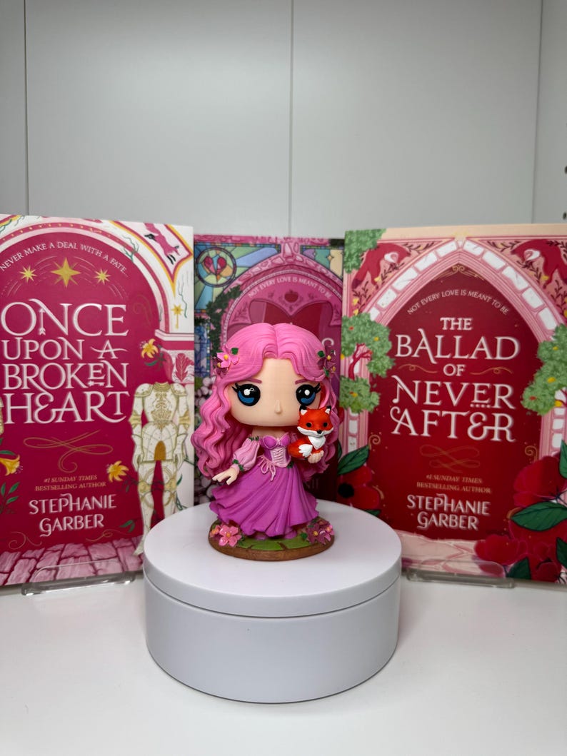 May include: A figurine of a girl with pink hair and a pink dress holding a small fox, placed in front of three books. The books have red and pink covers with the titles "Once Upon a Broken Heart" and "The Ballad of Never After" by Stephanie Garber.