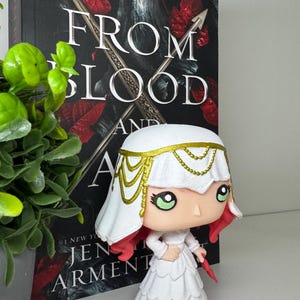 May include: A white figurine of a character from the book "From Blood and Ash" by Jennifer L. Armentrout. The figurine has a white dress, a white head covering with gold accents, and red hair. The book is in the background.