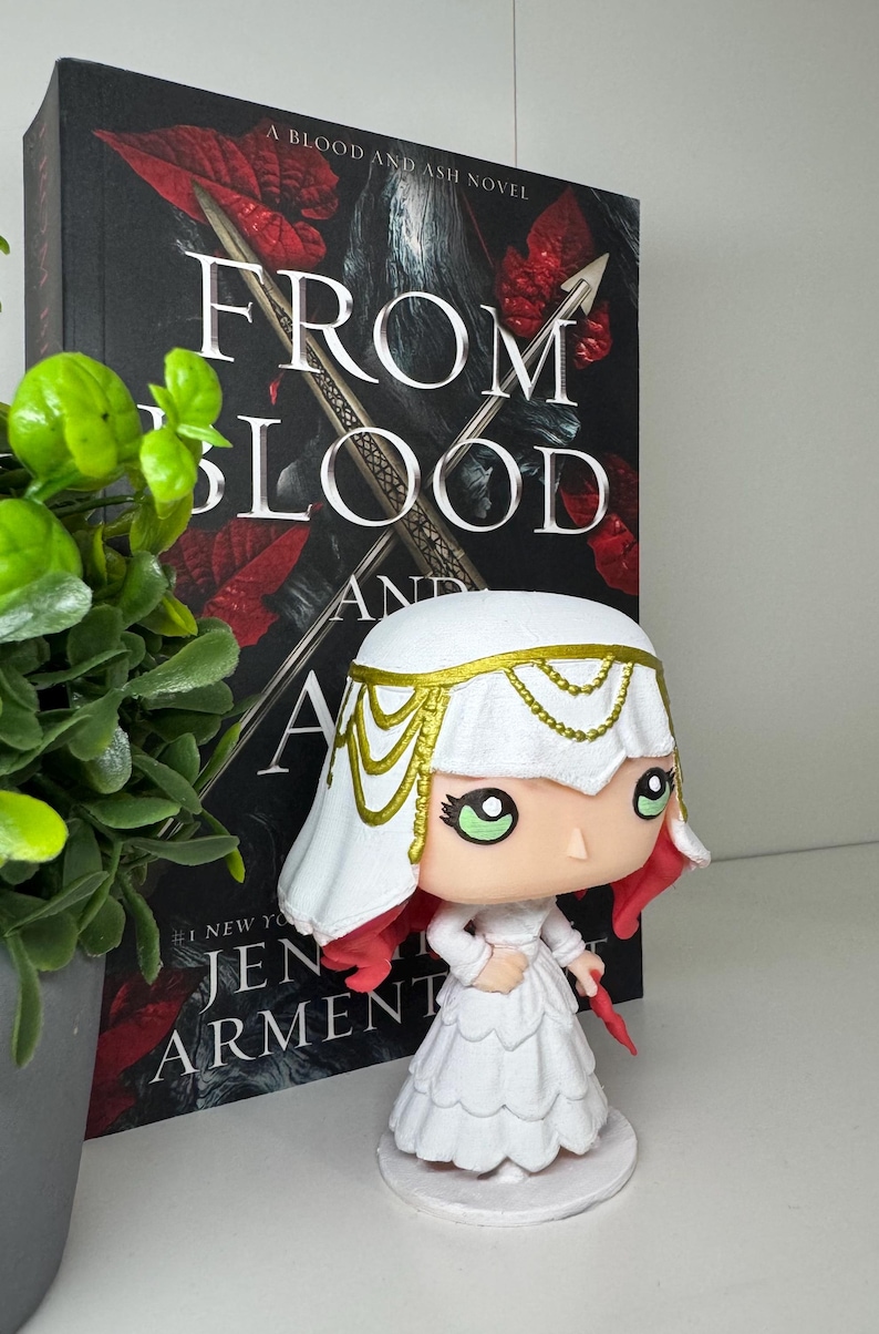 May include: A white figurine of a character from the book "From Blood and Ash" by Jennifer L. Armentrout. The figurine has a white dress, a white head covering with gold accents, and red hair. The book is in the background.