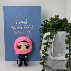 Pu&ograve; includere: Statuetta di una donna con un velo rosa e un abito nero, che tiene in mano una piccola pistola nera. Dietro la statuetta c'&egrave; un libro blu con testo dorato che recita "i spent my life folded between the". A destra c'&egrave; una fioriera in ceramica bianca con piante verdi sospese.