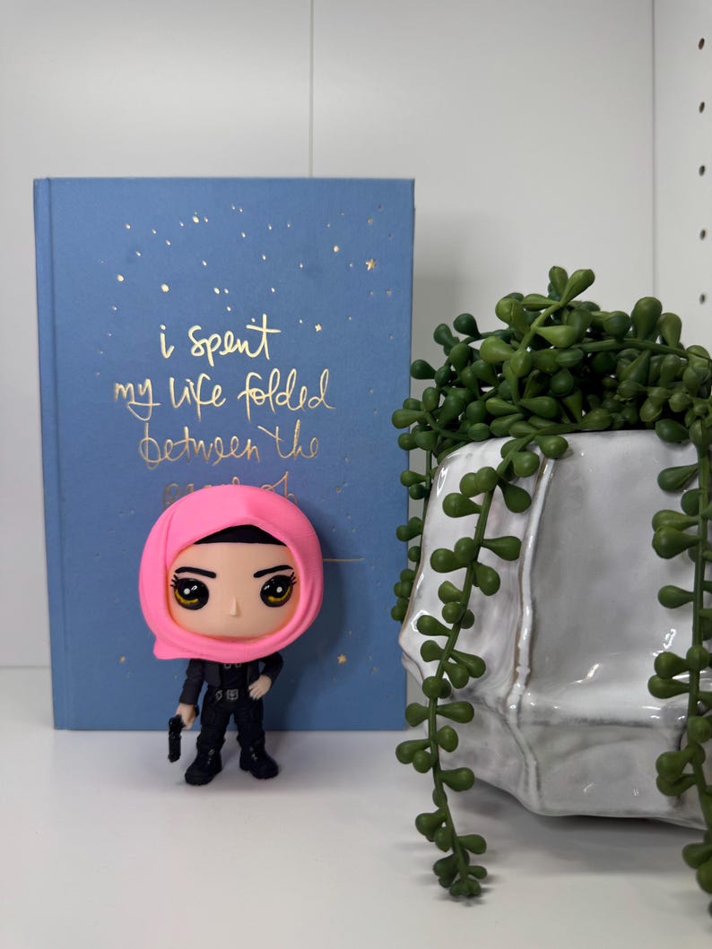 Pu&ograve; includere: Statuetta di una donna con un velo rosa e un abito nero, che tiene in mano una piccola pistola nera. Dietro la statuetta c'&egrave; un libro blu con testo dorato che recita "i spent my life folded between the". A destra c'&egrave; una fioriera in ceramica bianca con piante verdi sospese.