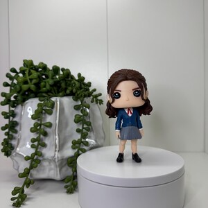 K&ouml;nnte beinhalten: Eine Vinylfigur einer Person in Schuluniform, mit blauem Blazer, grauem Rock und schwarzen Schuhen. Die Figur hat dunkles Haar und steht auf einer wei&szlig;en Plattform. Eine dekorative Pflanze in einem wei&szlig;en Topf befindet sich im Hintergrund.