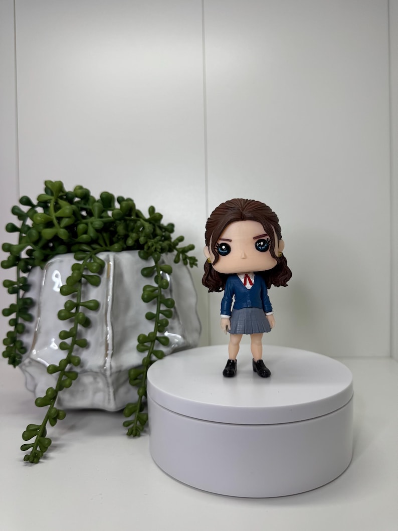 K&ouml;nnte beinhalten: Eine Vinylfigur einer Person in Schuluniform, mit blauem Blazer, grauem Rock und schwarzen Schuhen. Die Figur hat dunkles Haar und steht auf einer wei&szlig;en Plattform. Eine dekorative Pflanze in einem wei&szlig;en Topf befindet sich im Hintergrund.