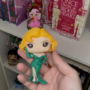 May include: A Funko Pop figure of a woman with blonde hair and a green dress. The figure is held in a hand, with books and other figurines on a shelf in the background. The book title "Once Upon a Broken Heart" is visible.