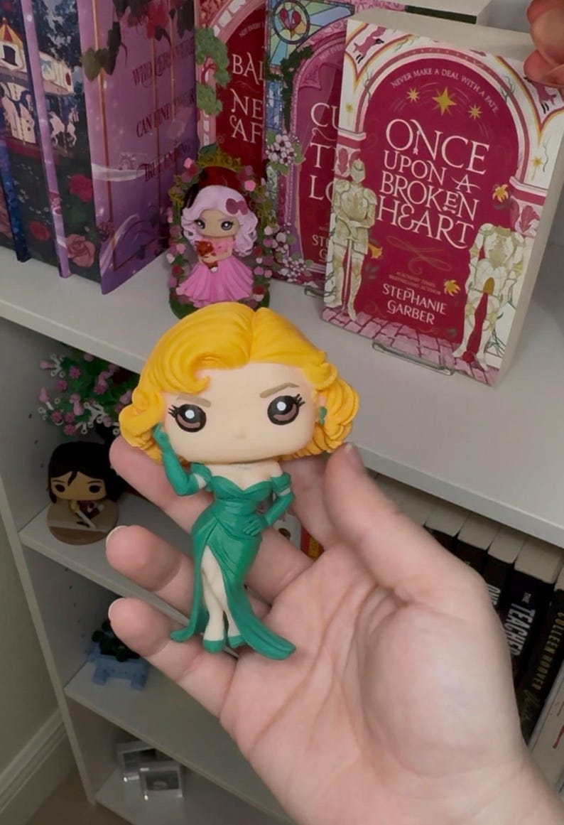 May include: A Funko Pop figure of a woman with blonde hair and a green dress. The figure is held in a hand, with books and other figurines on a shelf in the background. The book title "Once Upon a Broken Heart" is visible.