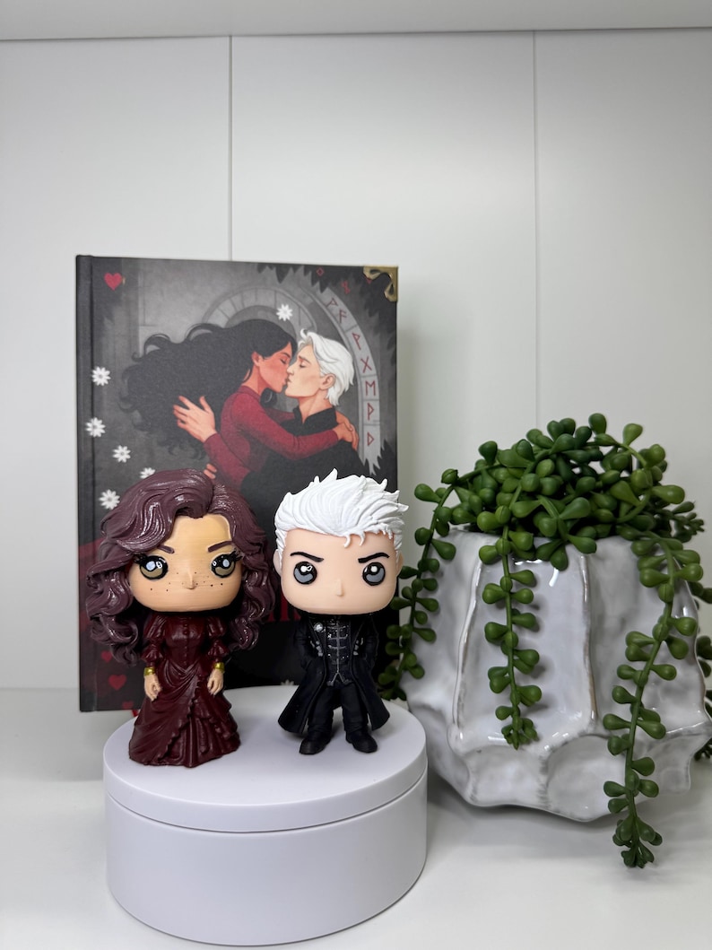 Może przedstawiać: Dwie figurki Funko Pop, jedna z bordowymi włosami i sukienką, druga z białymi włosami i czarnym strojem, stoją na białej platformie. Za nimi książka z całującą się parą na okładce. Po prawej stronie biała ceramiczna donica z zieloną rośliną.