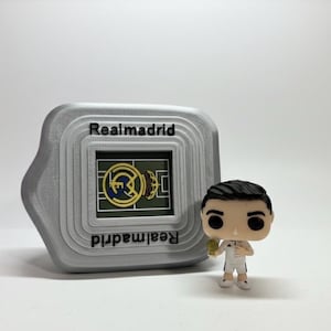 May include: A grey Realmadrid-themed digital device with the team's logo and name, alongside a figurine of a footballer in a white kit. The device has the words "Realmadrid" printed on it.