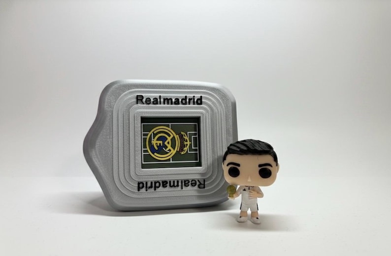 May include: A grey Realmadrid-themed digital device with the team's logo and name, alongside a figurine of a footballer in a white kit. The device has the words "Realmadrid" printed on it.