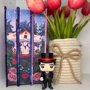 Puede incluir: Una exhibici&oacute;n con tres libros con ilustraciones de carrusel, una figura Funko Pop con traje y sombrero de copa, y un jarr&oacute;n con tulipanes rojos. La figura sostiene un bast&oacute;n.