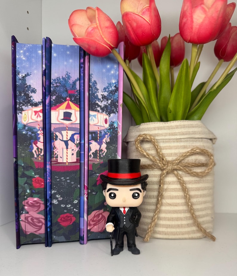 Puede incluir: Una exhibici&oacute;n con tres libros con ilustraciones de carrusel, una figura Funko Pop con traje y sombrero de copa, y un jarr&oacute;n con tulipanes rojos. La figura sostiene un bast&oacute;n.