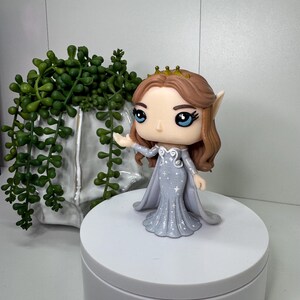 May include: A Funko Pop figure of a fantasy character. The figure has brown hair, blue eyes, and a gold crown. She wears a light gray dress with white star accents and a matching cape. The figure is on a white platform.
