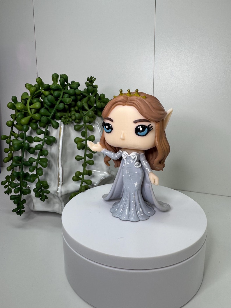May include: A Funko Pop figure of a fantasy character. The figure has brown hair, blue eyes, and a gold crown. She wears a light gray dress with white star accents and a matching cape. The figure is on a white platform.