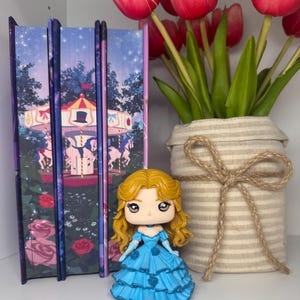 May include: A figurine of a woman in a blue dress with blonde hair stands in front of three decorative books. The books have a carousel scene on the cover. A vase with red tulips is to the right.