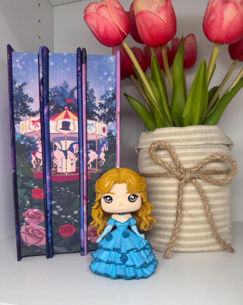 May include: A figurine of a woman in a blue dress with blonde hair stands in front of three decorative books. The books have a carousel scene on the cover. A vase with red tulips is to the right.