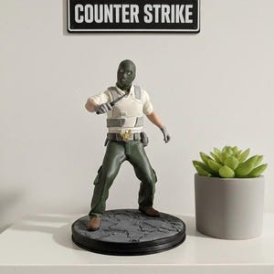 May include: A figurine of a character from Counter-Strike, wearing a green balaclava, vest, and pants, holding a knife. The figurine stands on a round base with a textured surface. A sign above reads "COUNTER STRIKE".