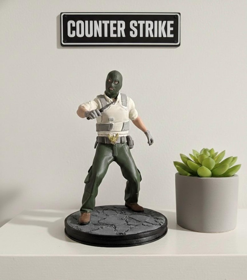 May include: A figurine of a character from Counter-Strike, wearing a green balaclava, vest, and pants, holding a knife. The figurine stands on a round base with a textured surface. A sign above reads "COUNTER STRIKE".