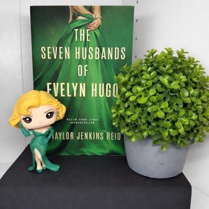 May include: A book titled "The Seven Husbands of Evelyn Hugo" with a green cover, a figurine of a woman in a green dress, and a small potted plant. The book is a New York Times bestseller by Taylor Jenkins Reid.