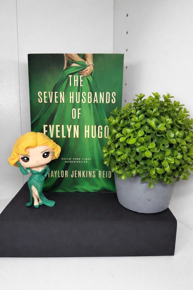 May include: A book titled "The Seven Husbands of Evelyn Hugo" with a green cover, a figurine of a woman in a green dress, and a small potted plant. The book is a New York Times bestseller by Taylor Jenkins Reid.