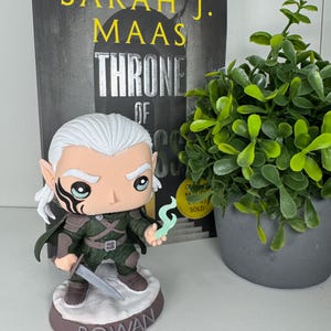 K&ouml;nnte beinhalten: Rowan Funko Pop Figur aus der Throne of Glass-Reihe, mit Schwert und magischem Wisp. Die Figur hat wei&szlig;es Haar und eine schwarze Gesichtsmarkierung. Auf dem Sockel steht "ROWAN". Ein Buch und eine kleine Topfpflanze sind im Hintergrund.