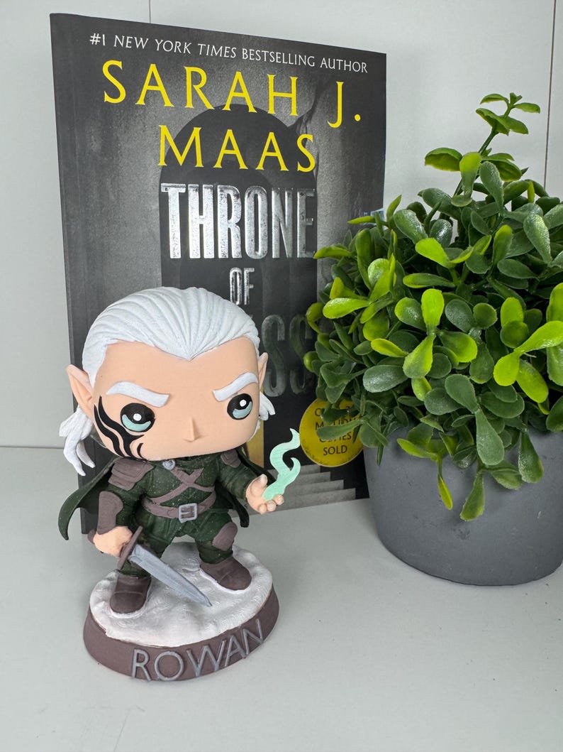 K&ouml;nnte beinhalten: Rowan Funko Pop Figur aus der Throne of Glass-Reihe, mit Schwert und magischem Wisp. Die Figur hat wei&szlig;es Haar und eine schwarze Gesichtsmarkierung. Auf dem Sockel steht "ROWAN". Ein Buch und eine kleine Topfpflanze sind im Hintergrund.