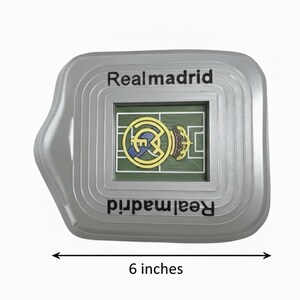 May include: A grey Real Madrid souvenir in the shape of a stadium, approximately 15 cm wide. It features a miniature football pitch with the team's logo. The text "Realmadrid" is printed on the top and bottom.