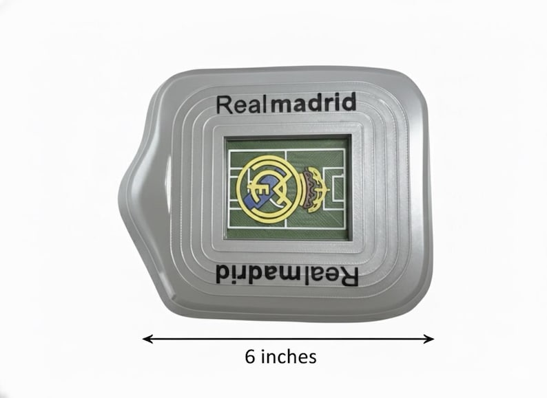 May include: A grey Real Madrid souvenir in the shape of a stadium, approximately 15 cm wide. It features a miniature football pitch with the team's logo. The text "Realmadrid" is printed on the top and bottom.