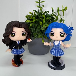 May include: Two character figurines with large eyes and detailed features. One has brown hair, a black blazer, and a blue plaid skirt. The other has blue hair and a blue plaid dress. Both are standing on decorative bases.