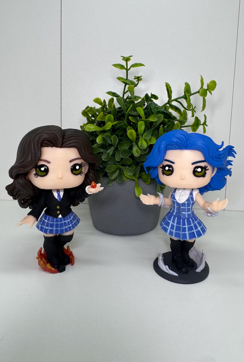 May include: Two character figurines with large eyes and detailed features. One has brown hair, a black blazer, and a blue plaid skirt. The other has blue hair and a blue plaid dress. Both are standing on decorative bases.
