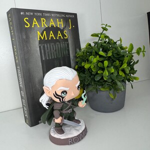 K&ouml;nnte beinhalten: Eine Figur von Rowan aus der Buchreihe "Throne of Glass" von Sarah J. Maas. Die Figur h&auml;lt ein Schwert und eine blaue Flamme. Das Buch mit dem Titel ist im Hintergrund zu sehen.