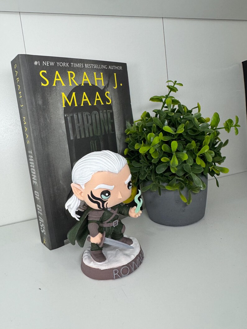 K&ouml;nnte beinhalten: Eine Figur von Rowan aus der Buchreihe "Throne of Glass" von Sarah J. Maas. Die Figur h&auml;lt ein Schwert und eine blaue Flamme. Das Buch mit dem Titel ist im Hintergrund zu sehen.