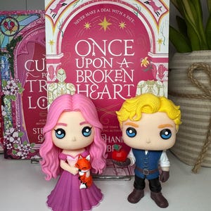 May include: Two Funko Pop figures of characters from "Once Upon a Broken Heart" stand in front of the book. The female figure has pink hair and a purple dress, holding a fox. The male figure has blonde hair and holds an apple. The book cover is visible.