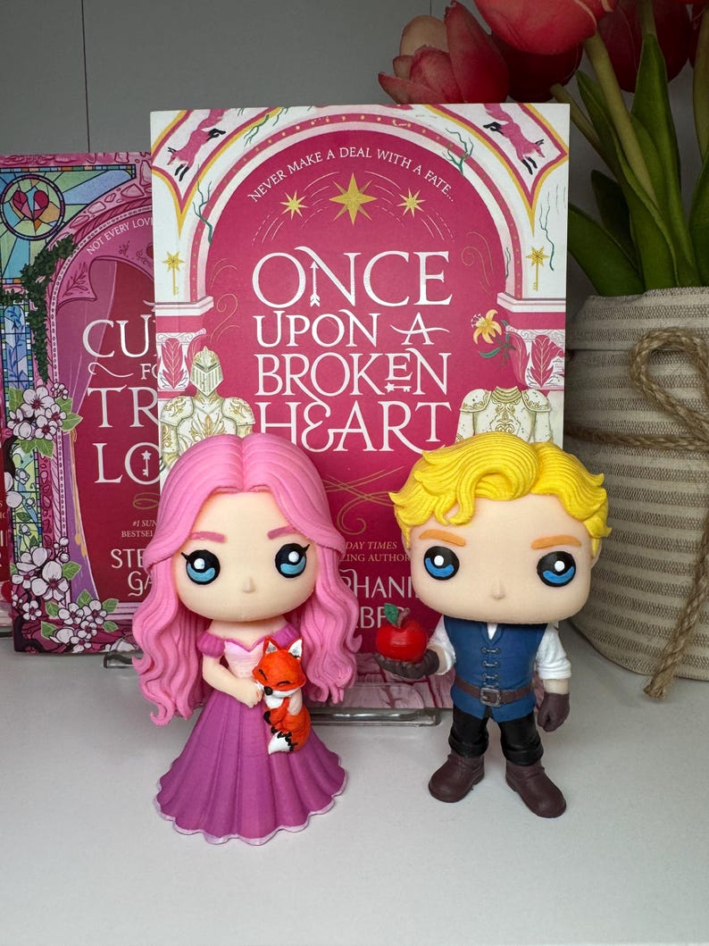 K&ouml;nnte beinhalten: Zwei Funko Pop-Figuren von Charakteren aus "Once Upon a Broken Heart" stehen vor dem Buch. Die weibliche Figur hat rosa Haare und ein lila Kleid und h&auml;lt einen Fuchs. Die m&auml;nnliche Figur hat blondes Haar und h&auml;lt einen Apfel. Das Buchcover ist sichtbar.