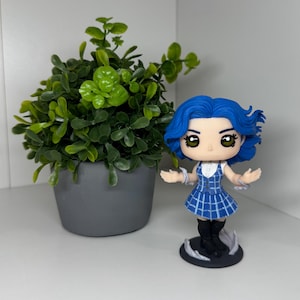 May include: A figurine of a character with blue hair and a blue and white plaid dress. The figurine has black boots and is standing on a black base with white accents. A small potted plant with green leaves is to the left.
